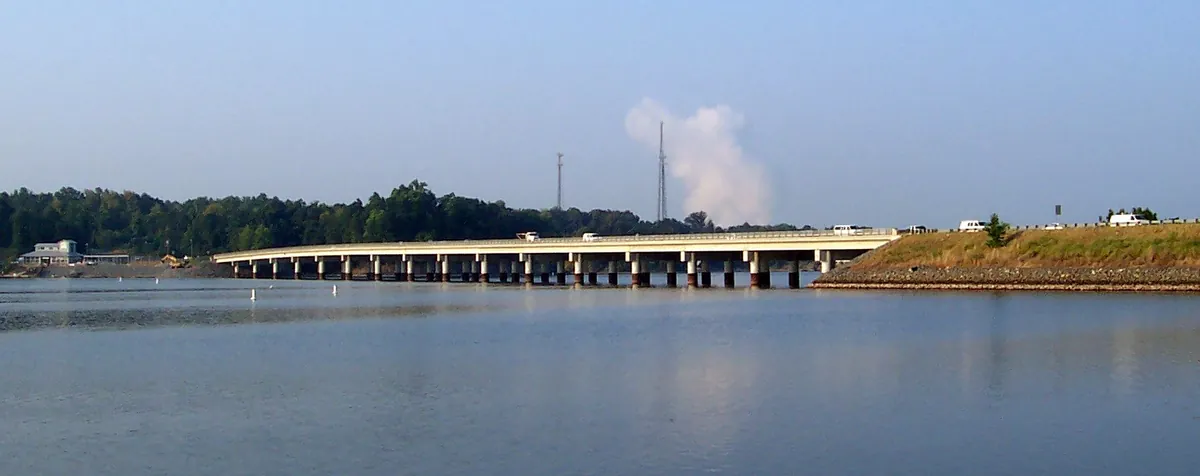 The long, low Buster Boyd Bridge crossing Lake Wylie on concrete piers, with a wooded shoreline and a white steam plume in the distance